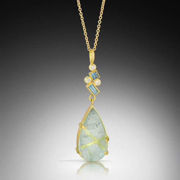 Photo of karin jacobson jewelry design pear shaped aquamarine and diamond necklace 18k yellow gold. The pear shaped rough aquamarine is in a hand fabricated 18k yellow gold setting with a cluster of faceted square and baguette aquamarines & round diamonds on the bail. Shown hanging on an 18k yellow gold cable chain. Shown on black background.