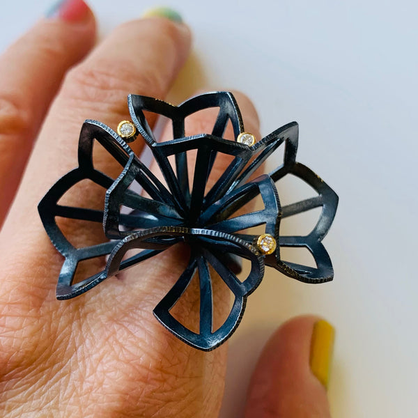 shows the top view of a large "origami" ring in oxidized sterling silver with 3 diamonds set in 18k gold. The origami looks kind of like a flower. Shown on a hand..