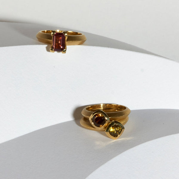 karin jacobson jewelry design solitaires in fairmined 18k yellow gold. Lower right shows caramel colored mali garnet ring on top of yellow mali garnet round sunburst cut bezel set ring with and upper left is pink tourmaline emerald cut ring.