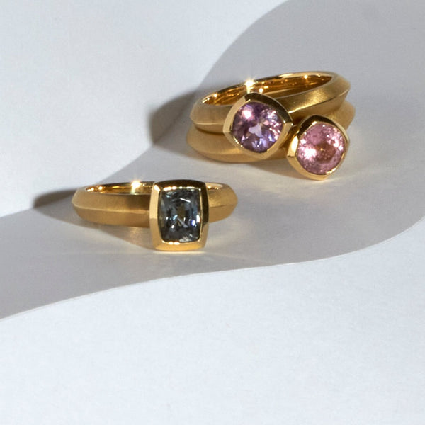 karin jacobson jewelry design - photo of three solitaires in fairmined 18k yellow gold. From left to right: gray spinel cushion cut bezel set ring; montana amethyst round cut bezel set ring; california pink tourmaline portuguese round cut bezel set ring.