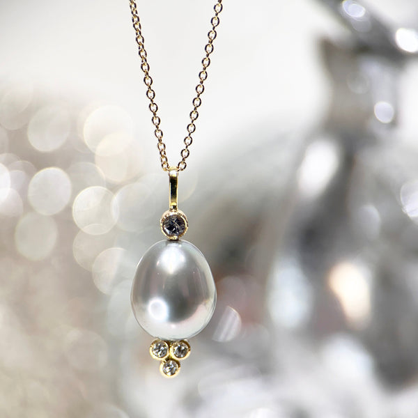 This photo shows an 18k yellow gold confetti necklace with a gray pearl - it has a 3mm gray sapphire on top and a cluster of three diamonds on the bottom - hanging from a gold cable chain, with a indistinct gray sparkly background.