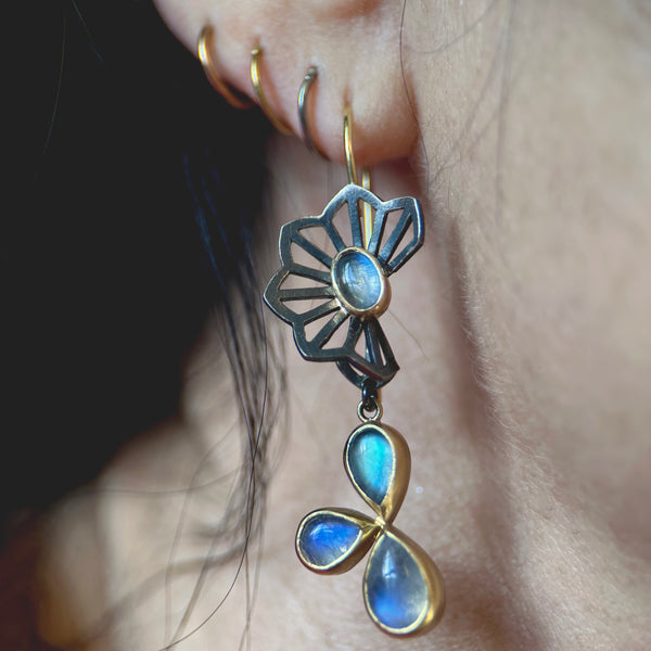 This photo shows a single earring on a model with an origami flower top in oxidized sterling silver with an oval moonstone in the center of the flower, and dangles with a cluster of three pear shaped moonstones, hanging from French wire.