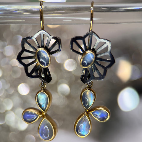 Shows a pair of earrings with origami flower tops in oxidized sterling silver with an oval moonstone in the center of the flower, and dangles with a cluster of three pear shaped moonstones, hanging from French wires - this version is in front of a sparkly background.