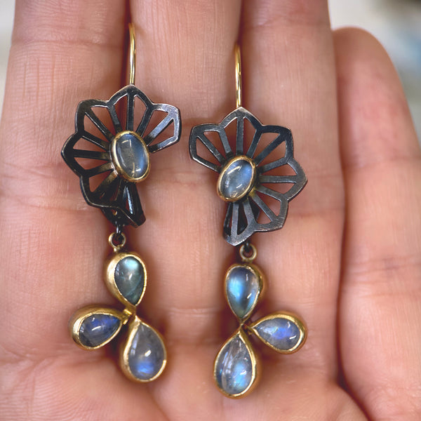 Shows a pair of earrings with origami flower tops in oxidized sterling silver with an oval moonstone in the center of the flower, and dangles with a cluster of three pear shaped moonstones, hanging from French wires - this version is shown held in a hand.