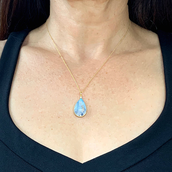 Photo of karin jacobson jewelry design pear shaped rainbow moonstone cabochon and diamond necklace in sterling silver, 22k & 18k yellow gold. It has a large pear moonstone that has inclusions and glows blue, with one diamond at the top of the pendant. Shown hanging from a gold chain and worn by a model.