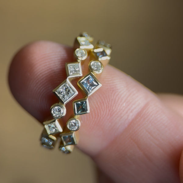 Close-up photo of two 18k yellow gold Confetti rings held between two fingers. One ring features scattered recycled diamonds, and the other combines blue sapphires with recycled diamonds. Both rings show mixed round and square stones arranged in the collection’s signature irregular pattern.