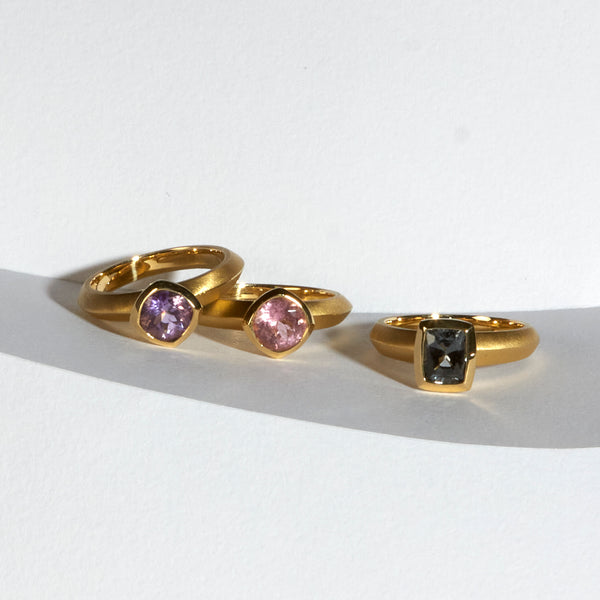 karin jacobson jewelry design - photo of three solitaires in fairmined 18k yellow gold. From left to right: montana amethyst round cut bezel set ring; california pink tourmaline portuguese round cut bezel set ring; and gray spinel cushion cut bezel set ring. 