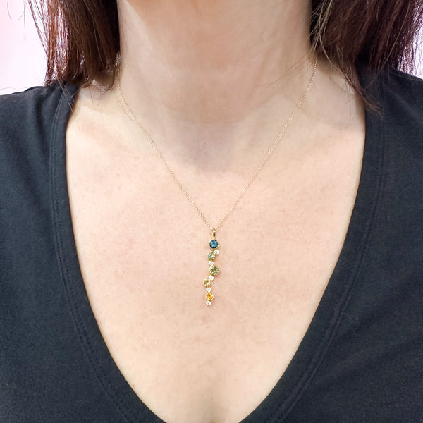 Shows an 18k yellow gold confetti necklace with various sizes of round Montana sapphires (in blue, teal, green, light yellow and dark yellow) and diamonds on a gold cable chain on a model.
