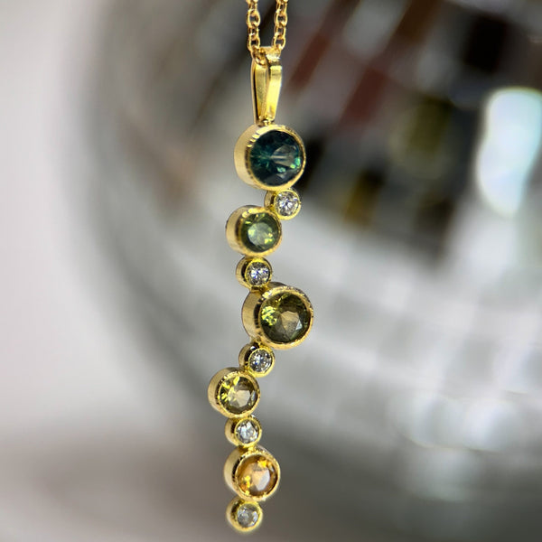 Shows an 18k yellow gold confetti necklace with various sizes of round Montana sapphires (in blue, teal, green, light yellow and dark yellow) and diamonds on a gold cable chain with an indistinct disco ball in the background.