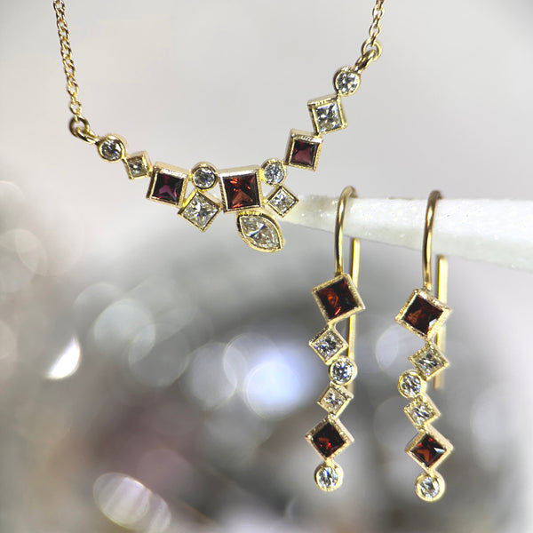 The photo shows 18k yellow gold confetti dangle earrings and a horizontal  confetti necklace with various square and round rusty red garnets and diamonds on a sparkly gray background.