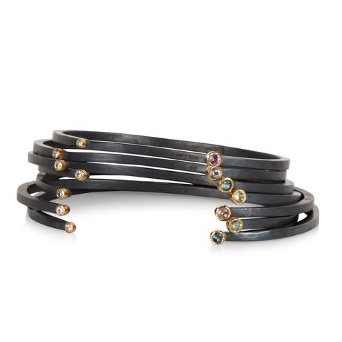 Stack of oxidized sterling silver cuff bracelets with a diamond on one end and a small round sapphire on the other, both set in 18k yellow gold.