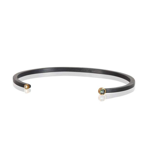 Oxidized sterling silver cuff bracelet with a diamond on one end and a small round teal sapphire on the other, both set in 18k yellow gold.