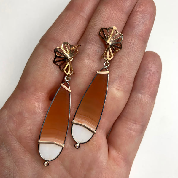 Pair of earrings with orange & white carnelian teardrop-shaped stones and 14k rose gold floral tops on a white background - this version shows the backs being held in a hand.