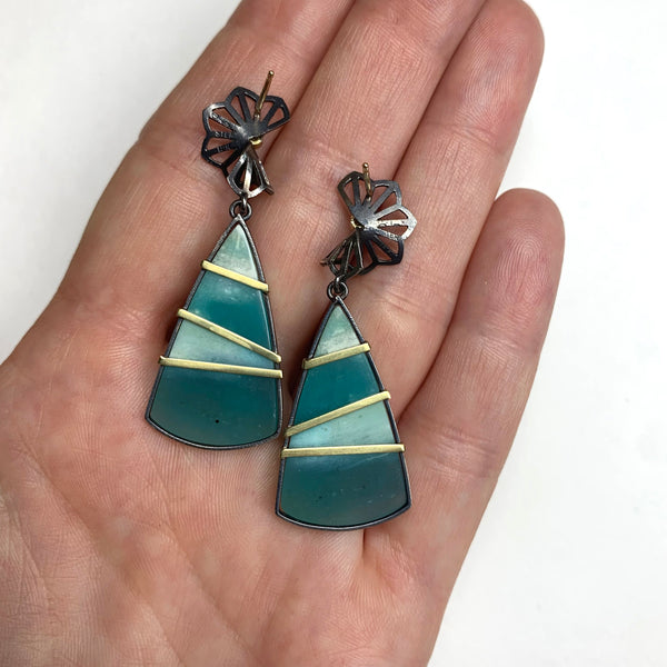  Photo of karin jacobson jewelry design opalized wood earrings with petite hyacinth fold tops. in sterling silver and 18k yellow gold with 2 diamonds. the opalized wood is triangle shaped with teal and white stripes. back sides shown in hand for scale